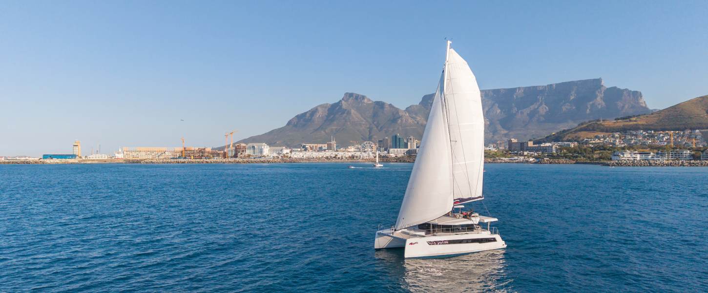 A sailing catamaran under sail with mainsail set, crew on the flybridge.