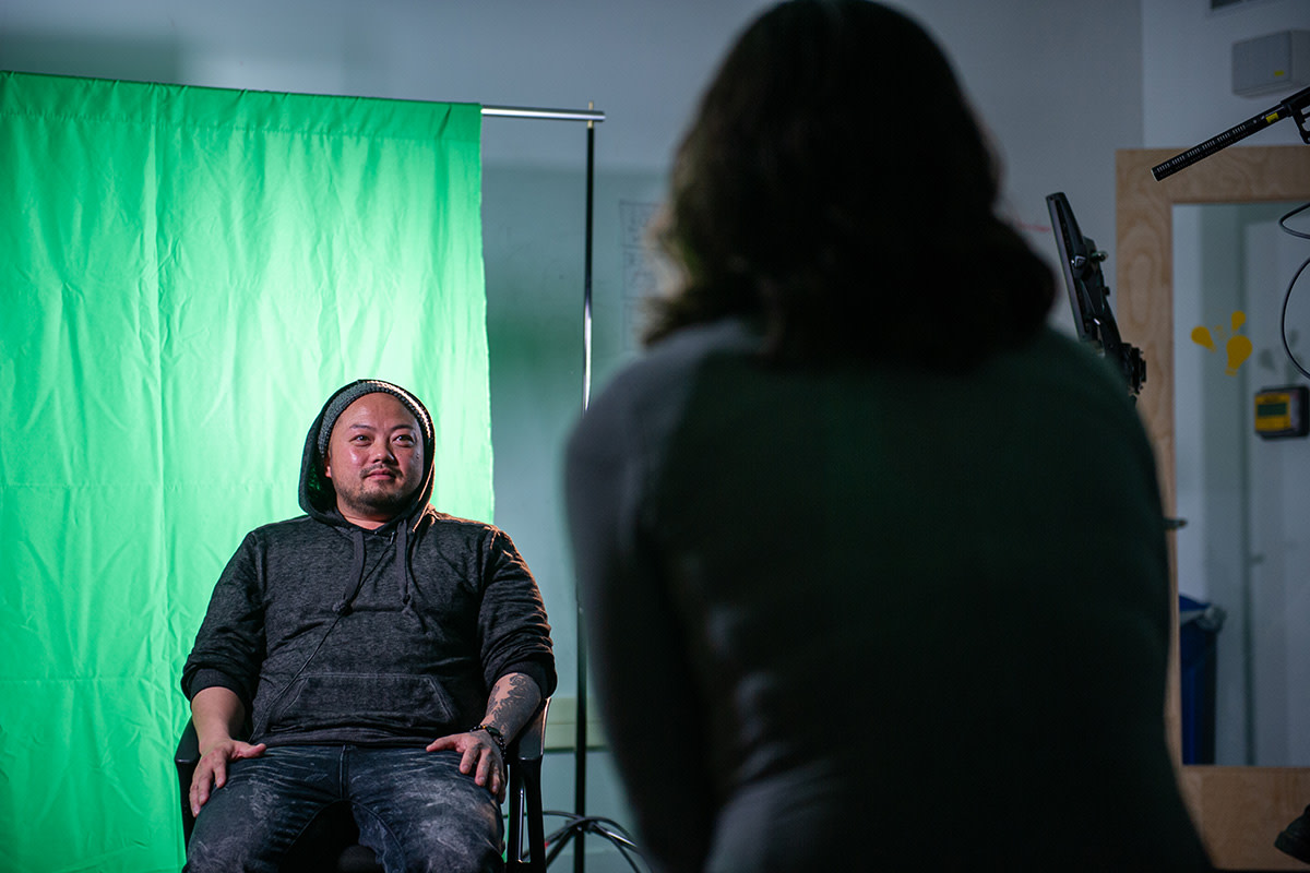 A seated individual in a hoodie in front of a green screen, with a camera person in the foreground