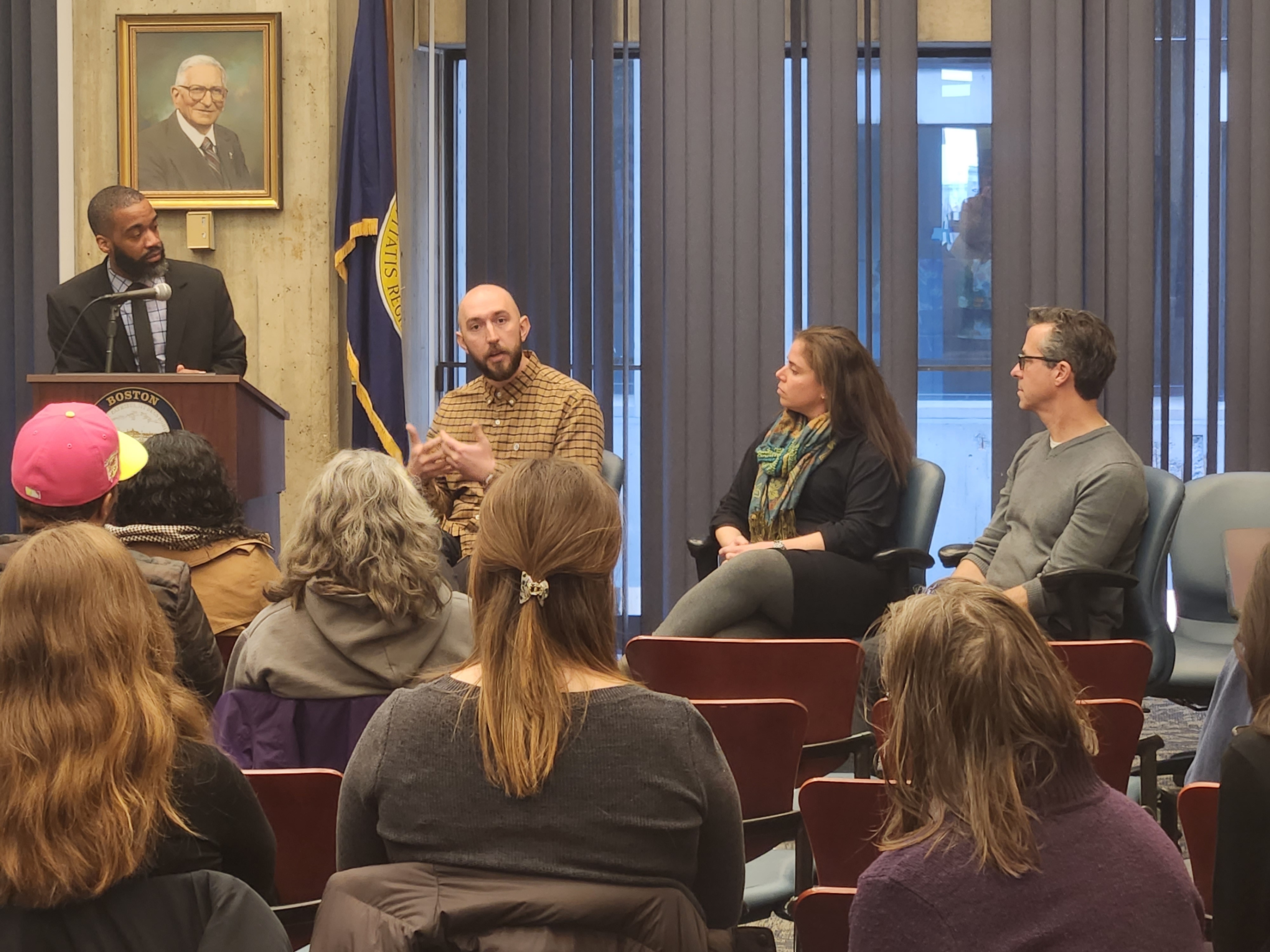 Three panelists seated in front of an audience at Boston City Hall, with moderator standing at a podium