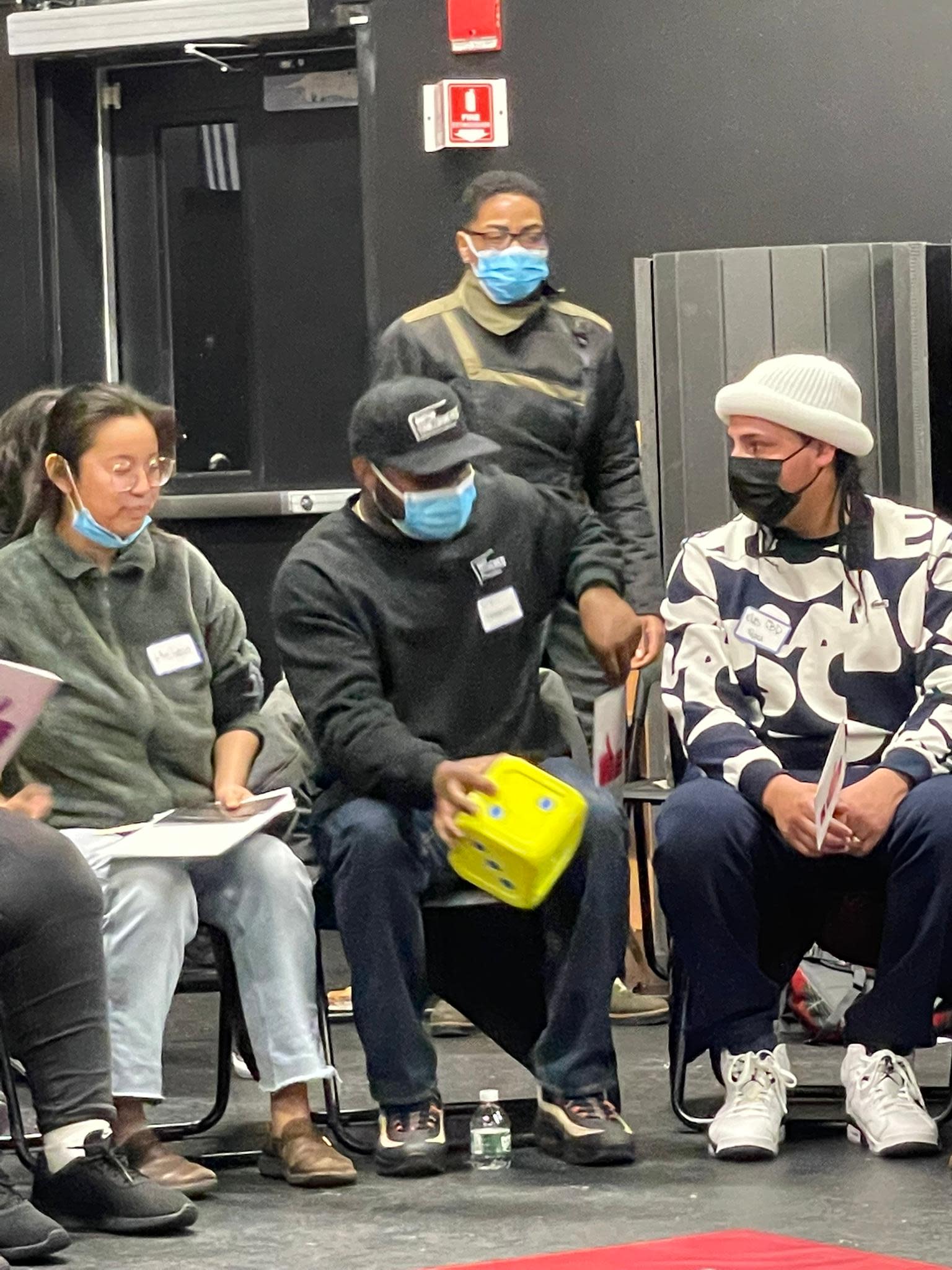 Three seated people in a blackbox theatre, one holding a large yellow die