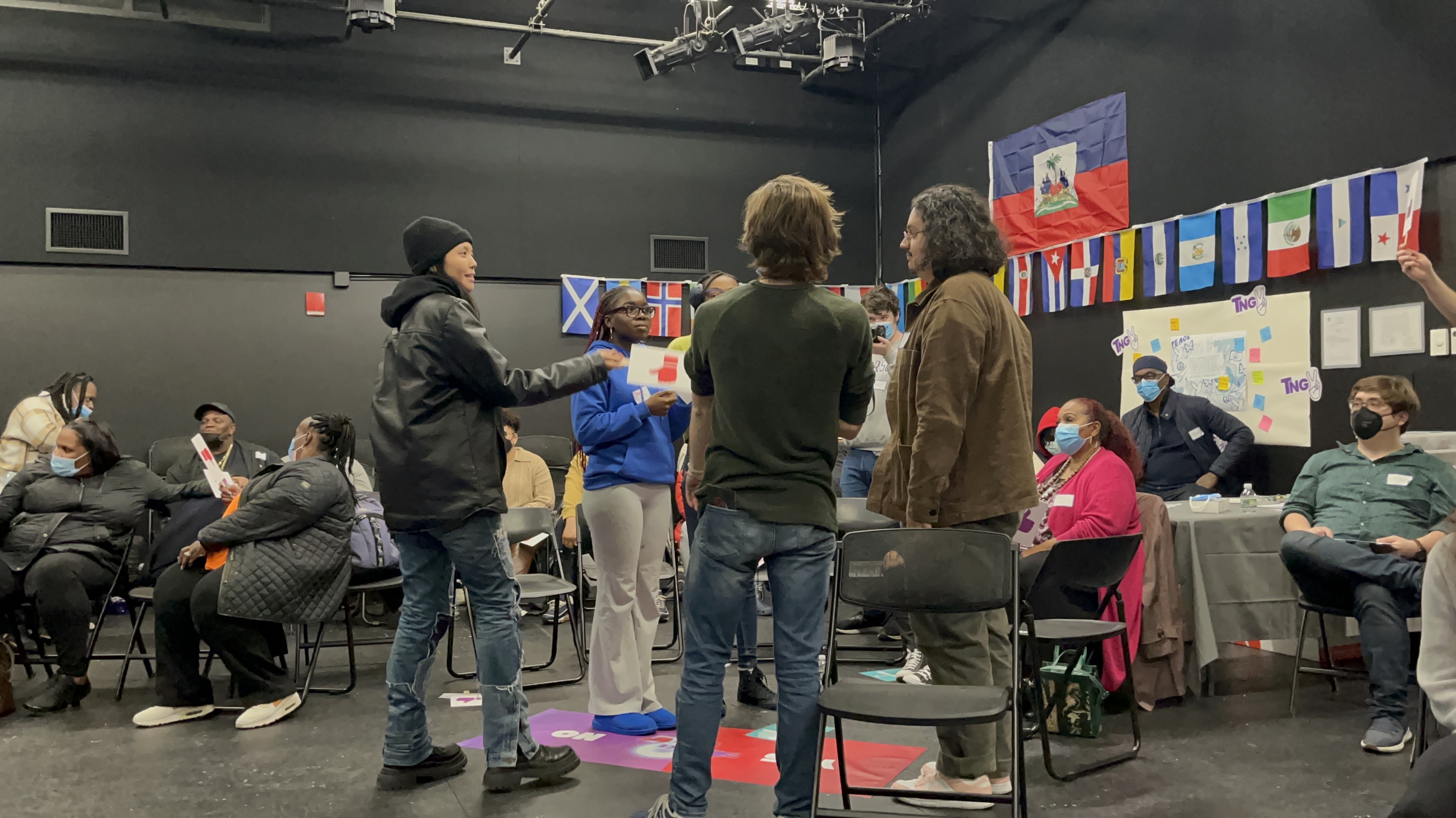 A number of people playing a role-playing game in a crowded black-box with flags on the wall