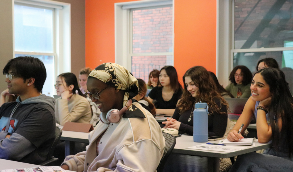 A group of smiling students seated at desks in a classroom with an orange accent wall