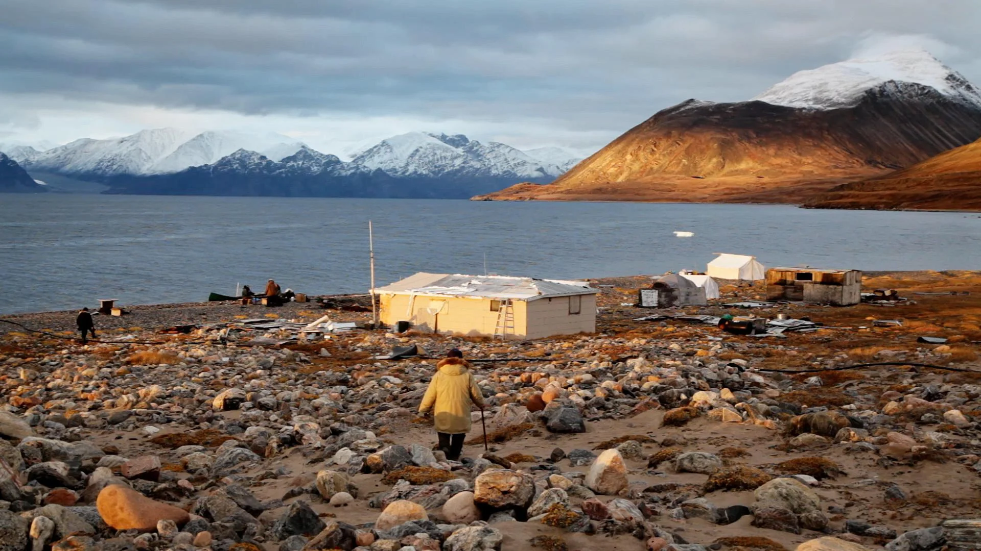 A person walking toward a white hut in front of a vast body of water and snowy mountains