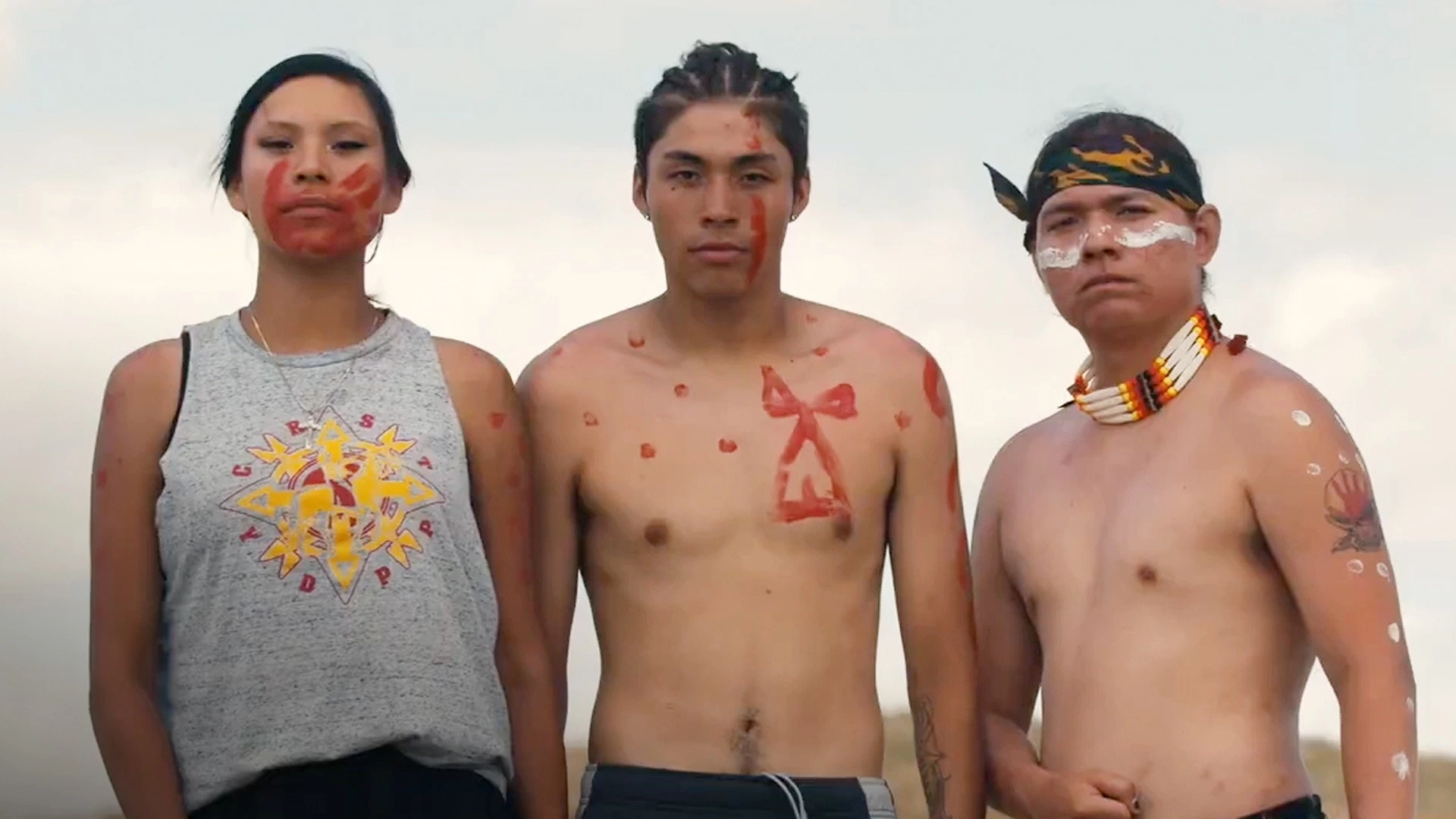 Three Native American people wearing some body paint and traditional jewelry, the person on the left wears a tank top with a Native graphic on it