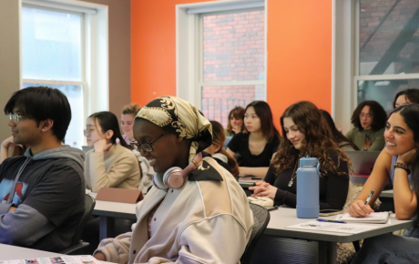 A group of smiling students seated at desks in a classroom with an orange accent wall