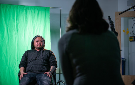 A seated individual in a hoodie in front of a green screen, with a camera person in the foreground