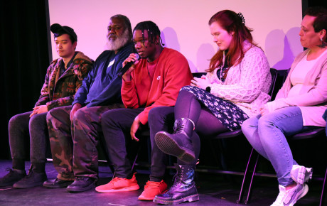 A gathering of five individuals seated on stage, one of whom is speaking into a microphone