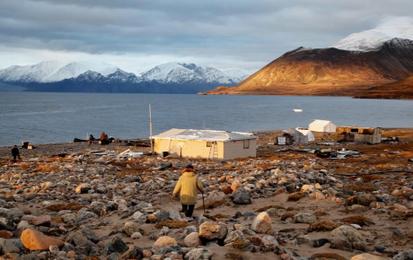 A person walking toward a white hut in front of a vast body of water and snowy mountains