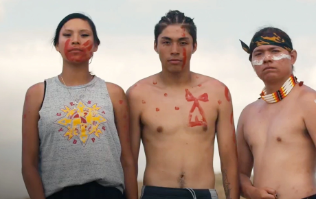 Three Native American people wearing some body paint and traditional jewelry, the person on the left wears a tank top with a Native graphic on it