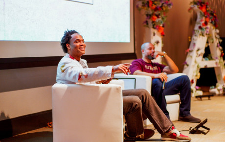 Two individuals seated on a stage, looking at the audience, with a projector screen behind them