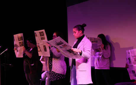 Four individuals performing on a stage, holding newspapers
