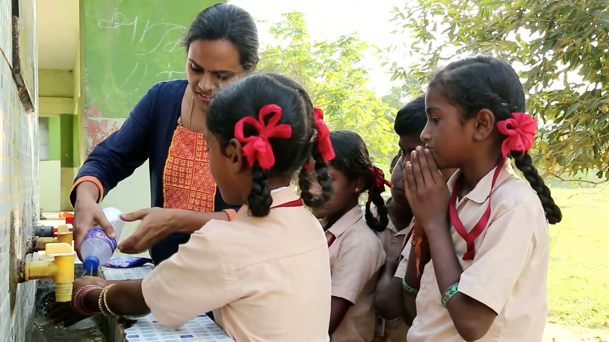 Students gather around a teacher to wash their hands with a soapy bottle.