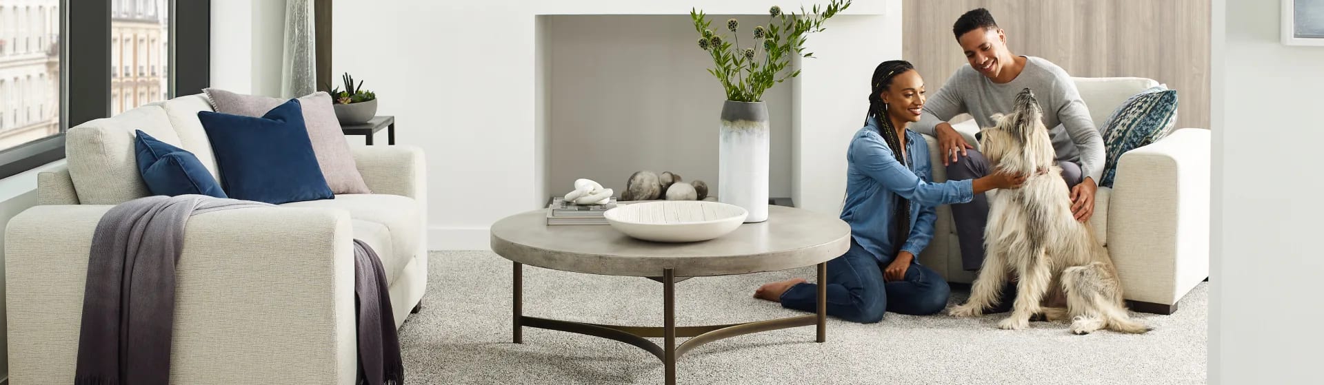 A couple sitting on the floor of a modern living room, playing with their dog.