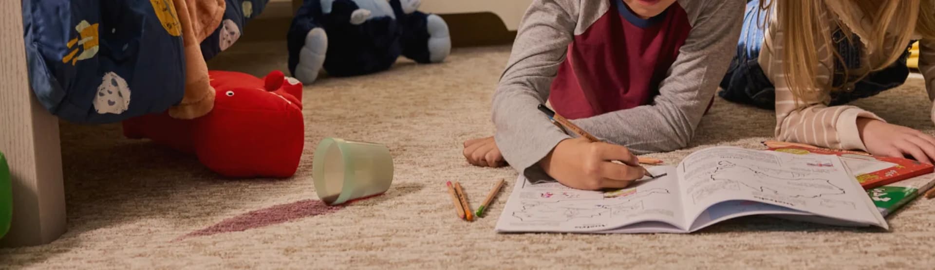 Children drawing on paper spread out across a soft carpet with a spilled drink nearby.