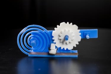 The 3-D printed gears (in white) and spring (blue spiral) toggle a switch (white box with grey surface) made of conductive plastic. The switch changes the reflective state of a 3-D printed antenna (gray strip) to convey useful data to a WiFi receiver. The shape of the gears and the speed at which they move encode the digital data. (Image courtesy of Mark Stone/University of Washington.)