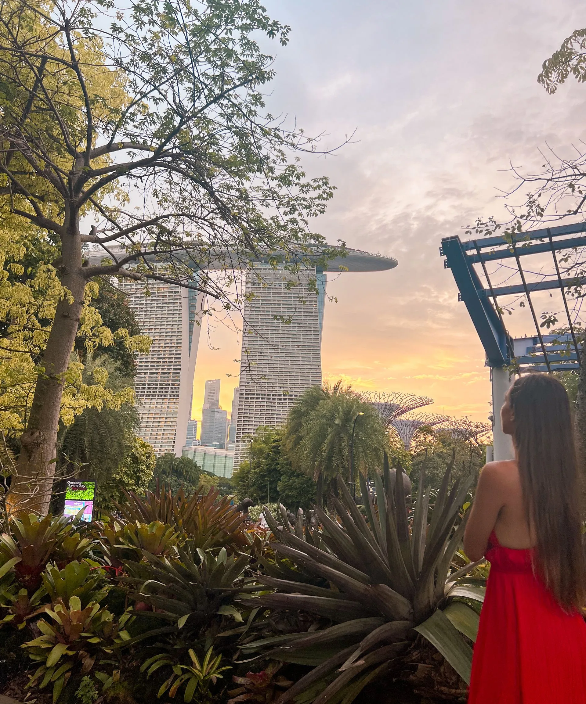 Frau im roten Kleid blickt auf Marina Bay Sands und Gardens by the Bay bei Sonnenuntergang, Singapur