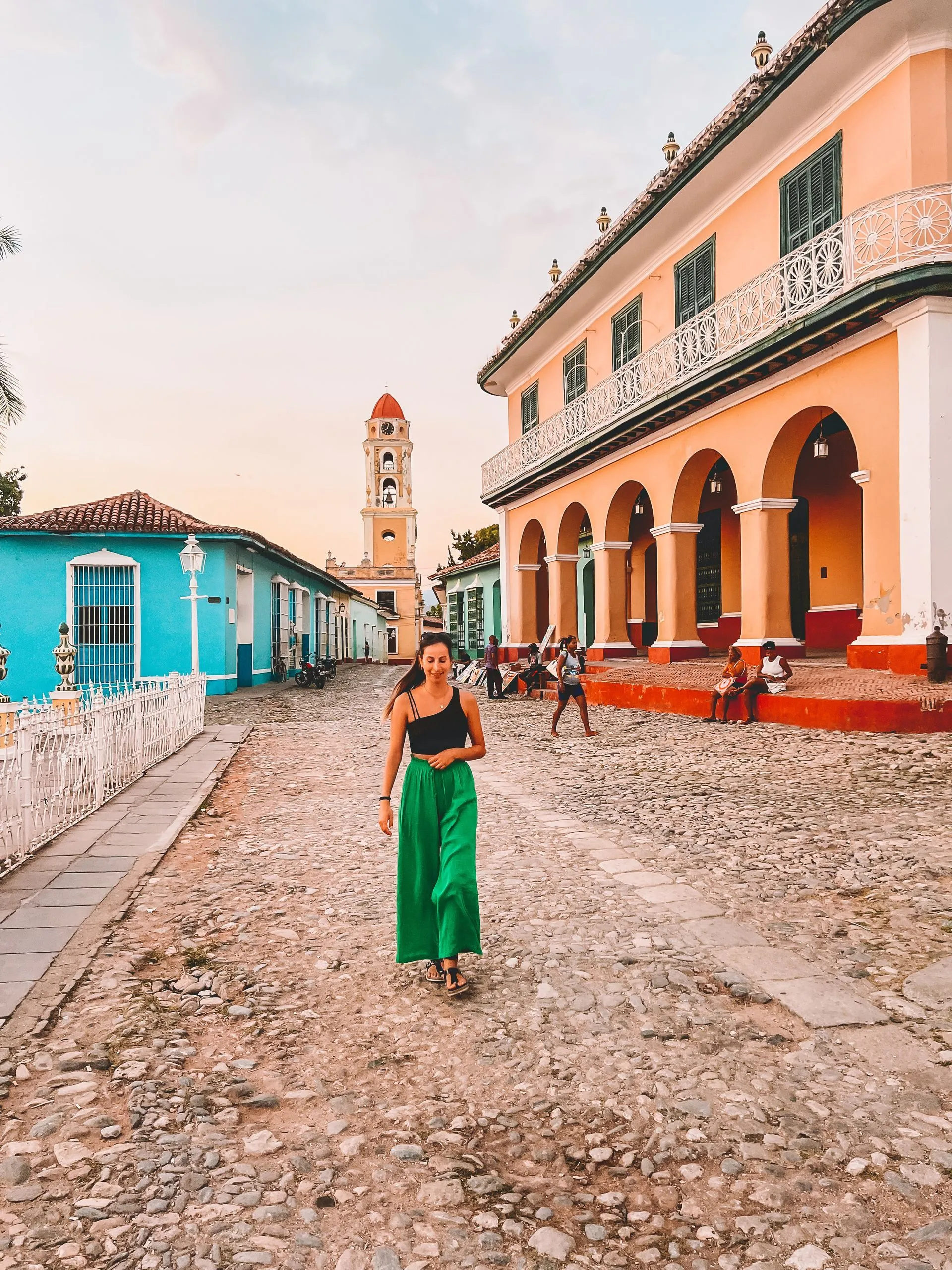 Frau in grünen Palazzo-Hosen spaziert über kopfsteinbepflastertem Platz in Trinidad, Kuba