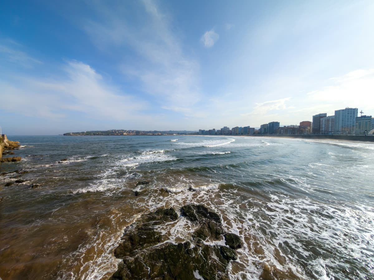 Playa de San Lorenzo en Gijón con olas y el skyline de la ciudad al fondo
