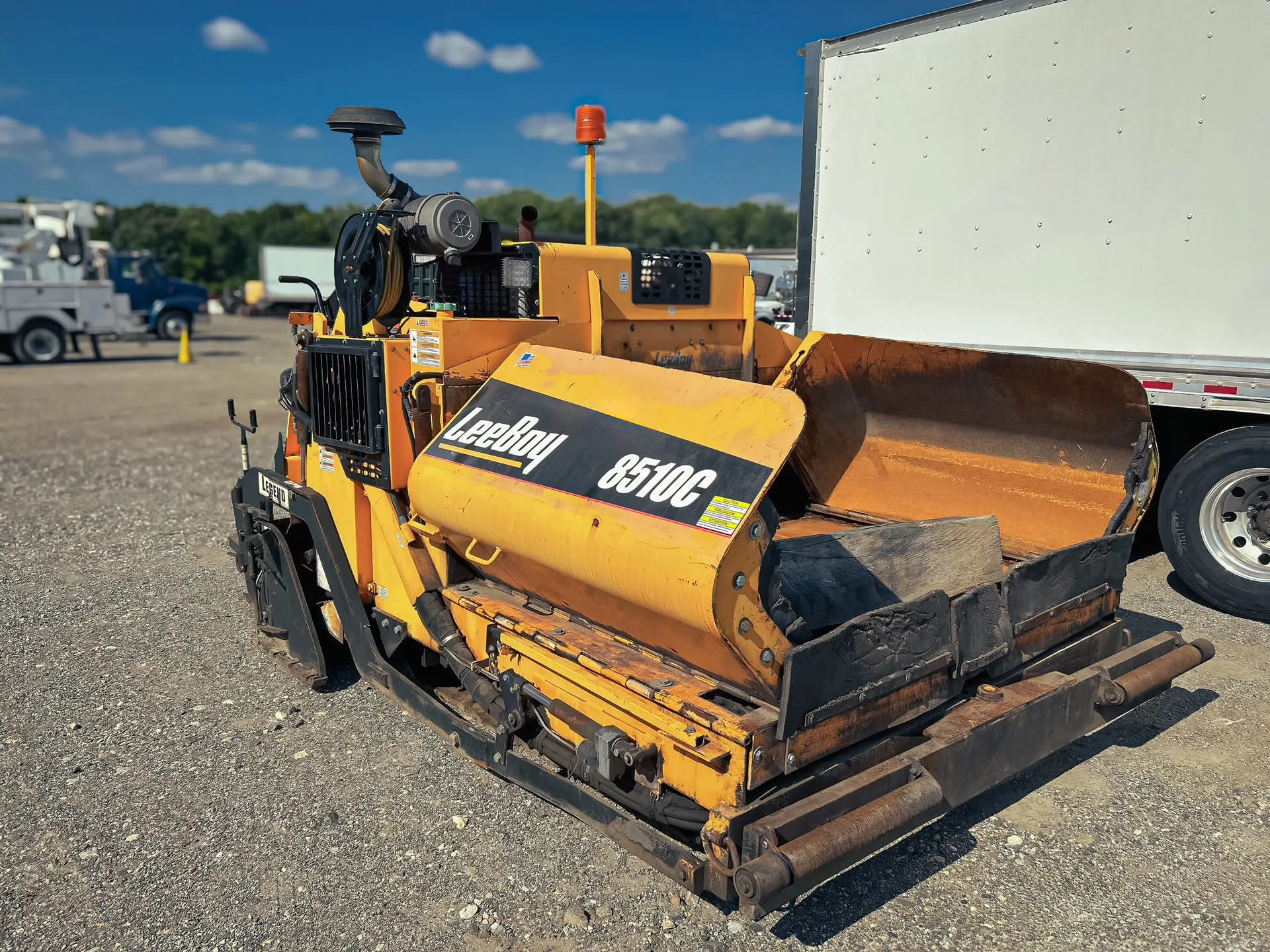 LeeBoy 8510C  A yellow LeeBoy 8510C track asphalt paver is parked on a gravel lot. It features dual controls, and is positioned next to a white truck trailer. The machine is designed for paving asphalt, with prominent branding on its side.