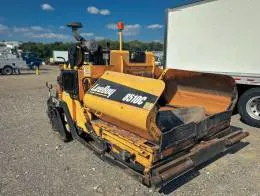 LeeBoy 8510C  A yellow LeeBoy 8510C track asphalt paver is parked on a gravel lot. It features dual controls, and is positioned next to a white truck trailer. The machine is designed for paving asphalt, with prominent branding on its side.