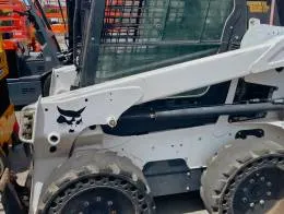 Bobcat S550 Skid_Steers A side view of a Bobcat S550 skid steer loader with a white and black color scheme. The machine has protective mesh windows, and solid rubber tires with a unique tread pattern featuring holes around the edges. The loader arms are in a lowered position.