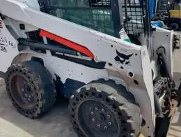 Bobcat S550 Skid_Steers A close-up side view of a Bobcat S550 skid steer loader. The loader's white body features a distinctive black logo, and the vehicle is equipped with solid rubber tires with multiple circular holes around the rim. The cab has metal protective grates on the windows.