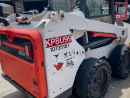 Bobcat S550 Skid_Steers  A rear view of a Bobcat S550 skid steer loader. The machine has a white body with a red rear panel and solid tires with circular holes along the edges. The loader arms are visible, along with identification markings on the side of the machine.