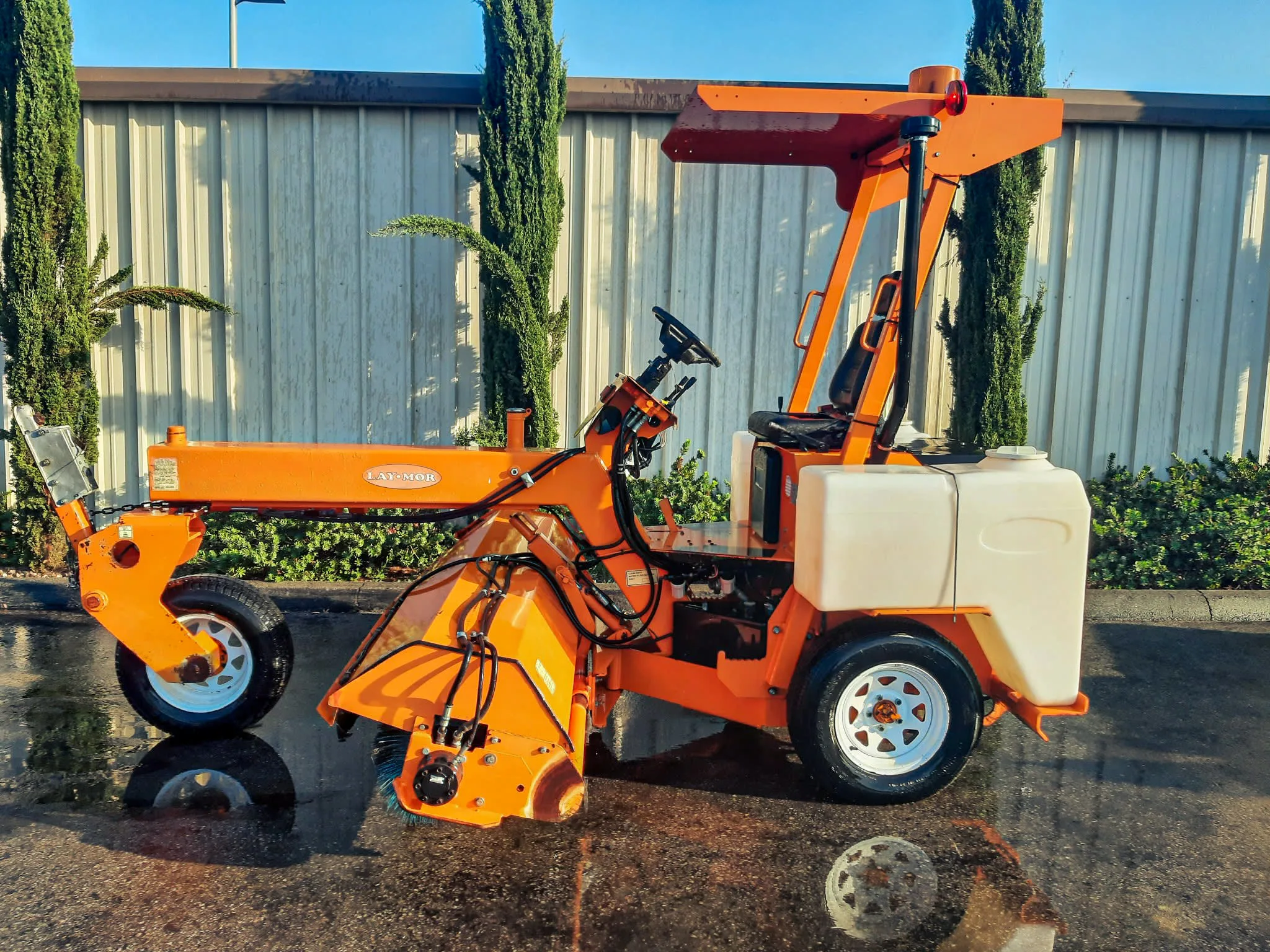Laymor 8HC Sweepers A bright orange Lay-Mor 8HC Sweeper machine is parked outdoors, in front of a office building with a row of tall trees in the background. The machine has a canopy, two white water tanks on the rear sides, and a long sweeping mechanism at the front. The reflection of the sweeper is visible on the wet pavement beneath it, highlighting its freshly washed appearance.