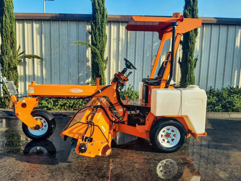 Laymor 8HC Sweepers A bright orange Lay-Mor 8HC Sweeper machine is parked outdoors, in front of a office building with a row of tall trees in the background. The machine has a canopy, two white water tanks on the rear sides, and a long sweeping mechanism at the front. The reflection of the sweeper is visible on the wet pavement beneath it, highlighting its freshly washed appearance.