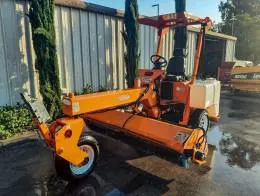 Laymor 8HC Sweepers A bright orange Lay-Mor 8HC Sweeper is shown parked outdoors in front of office building, angled to display the front sweeping mechanism. The sweeper has a canopy above the driver's seat and two white water tanks on the rear. The machine’s large front brushes and compact design are visible, with reflections of the equipment on the wet pavement below.Trees, and other machinery, including a LeeBoy 8515C, are in the background. The scene is lit by the afternoon sun, casting long shadows.