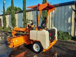 Laymor 8HC Sweepers A bright orange Lay-Mor 8HC Sweeper machine is parked in front of office building, viewed from a rear angle. The sweeper has a canopy over the operator's seat and two large white water tanks mounted on the back. The rear of the machine has black and red components, while the sweeping mechanism at the front is visible. The machine sits on clean pavement with reflections of the sweeper and nearby trees in the wet ground. Tall trees and electrical lines are seen in the background under a clear blue sky.