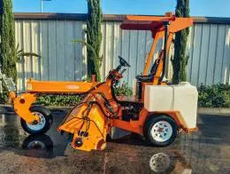 Laymor 8HC Sweepers A bright orange Lay-Mor 8HC Sweeper machine is parked outdoors, in front of a office building with a row of tall trees in the background. The machine has a canopy, two white water tanks on the rear sides, and a long sweeping mechanism at the front. The reflection of the sweeper is visible on the wet pavement beneath it, highlighting its freshly washed appearance.