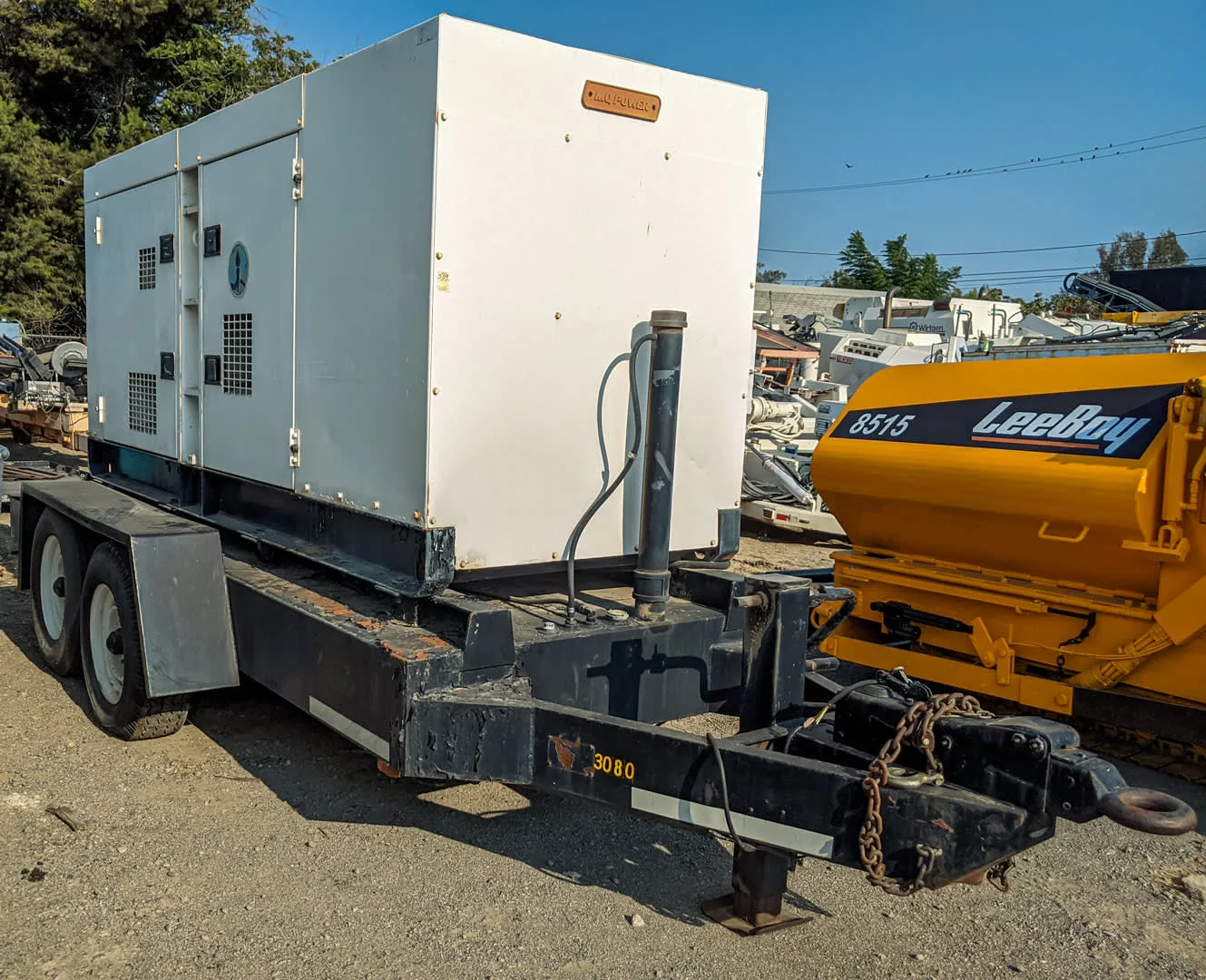 MultiQuip DCA-180SSK Generators The image shows a towable Multiquip 180KVA generator mounted on a heavy-duty dual-axle trailer. The generator housing is white, with ventilation grilles and latches on the sides for access. The trailer has a black frame and it includes a hitch setup for towing, complete with a chain and heavy-duty hook. In the background, a yellow LeeBoy 8515 machine is visible, along with other industrial equipment. The scene appears to be in an outdoor yard or equipment lot.
