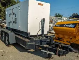 MultiQuip DCA-180SSK Generators The image shows a towable Multiquip 180KVA generator mounted on a heavy-duty dual-axle trailer. The generator housing is white, with ventilation grilles and latches on the sides for access. The trailer has a black frame and it includes a hitch setup for towing, complete with a chain and heavy-duty hook. In the background, a yellow LeeBoy 8515 machine is visible, along with other industrial equipment. The scene appears to be in an outdoor yard or equipment lot.