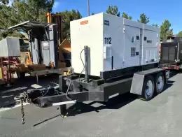 MultiQuip DCA-180SSK Generators The image shows a towable Multiquip 180KVA generator mounted on a heavy-duty dual-axle trailer. The generator housing is white, with ventilation grilles and latches on the sides for access. The trailer has a black frame and it includes a hitch setup for towing, complete with a chain and heavy-duty hook. In the background, a yellow LeeBoy 8515 machine is visible, along with other industrial equipment. The scene appears to be in an outdoor yard or equipment lot.