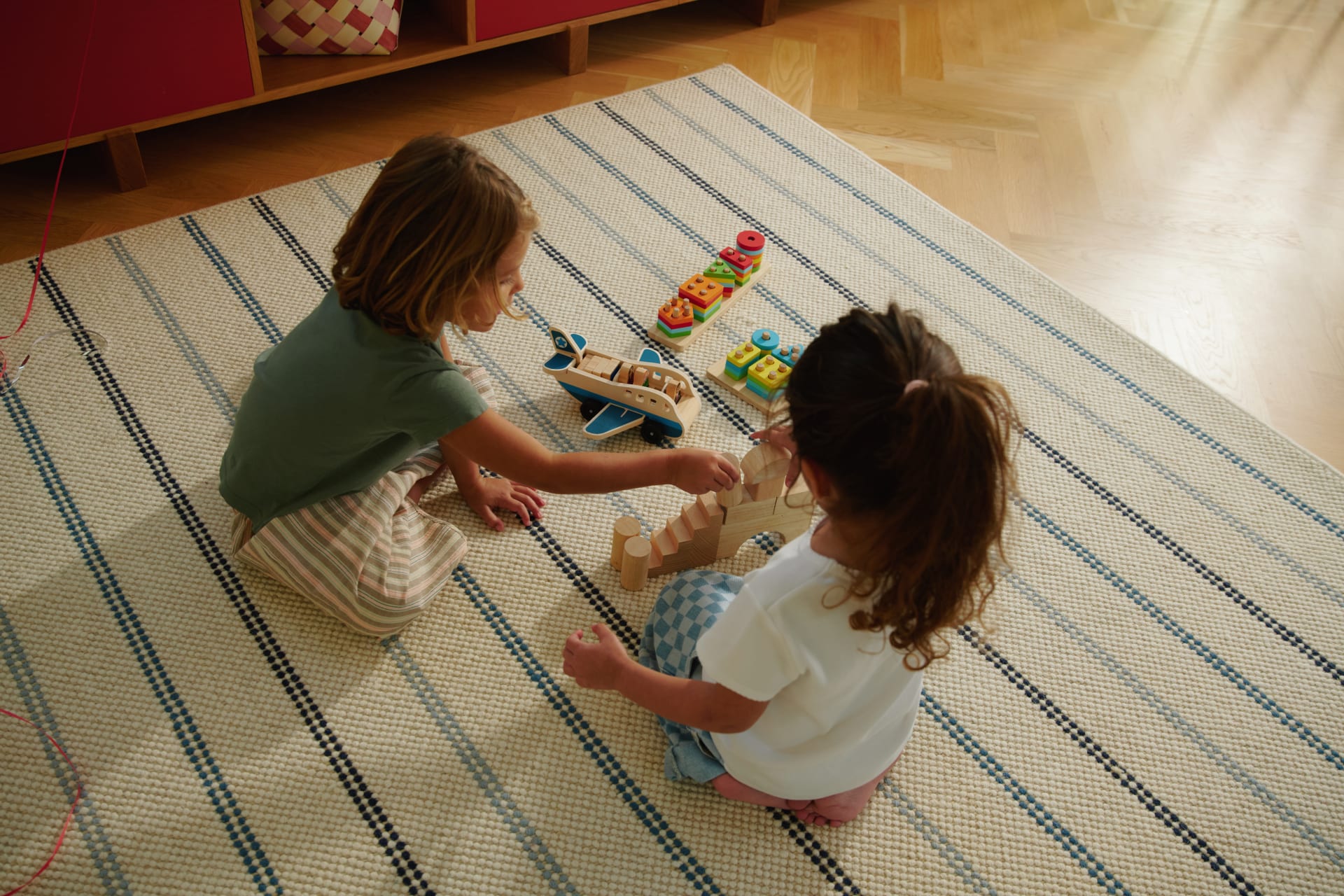 Two children playing with wooden blocks and toy vehicles on a striped carpet in a cozy room.