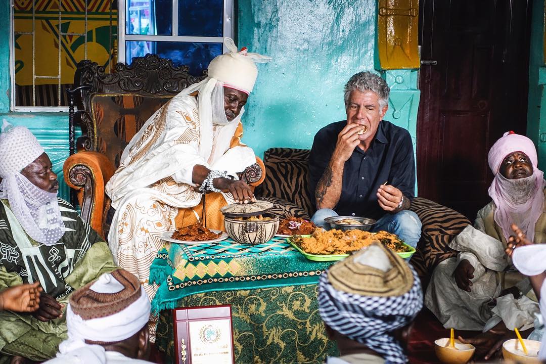 Anthony Bourdain eating lunch with four ornately dressed tribe chiefs.