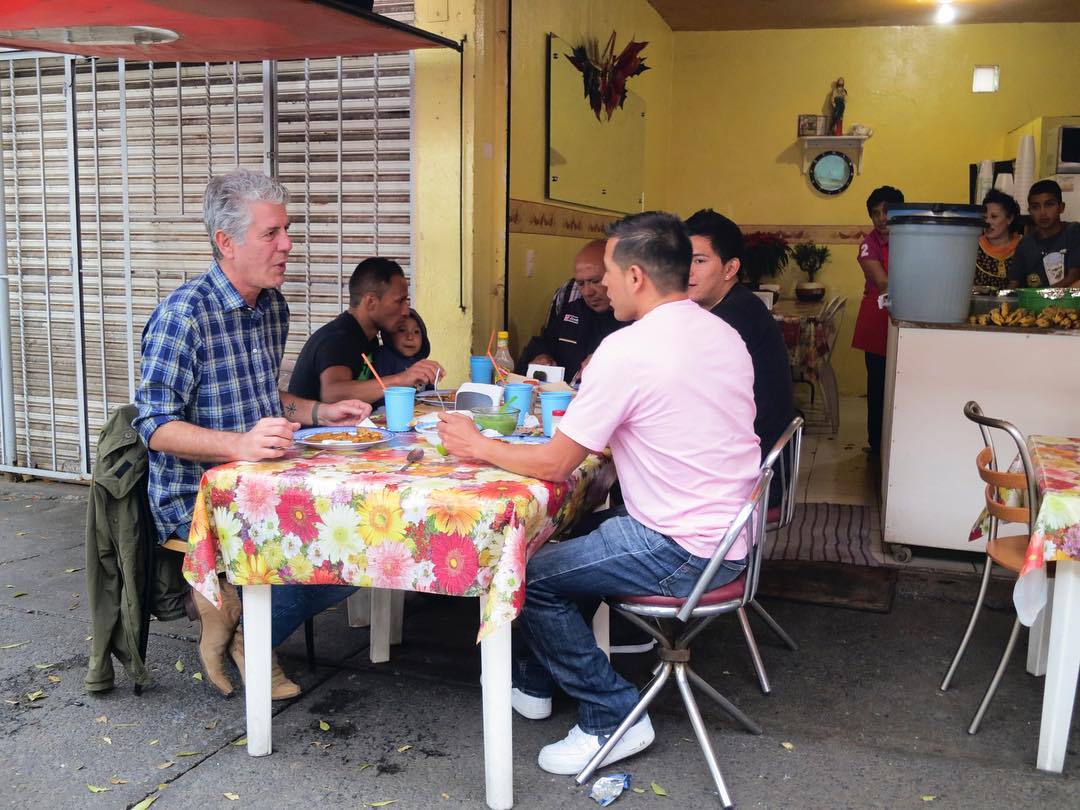 Anthony Bourdain eating with two fixers in a table set on a sidewalk.