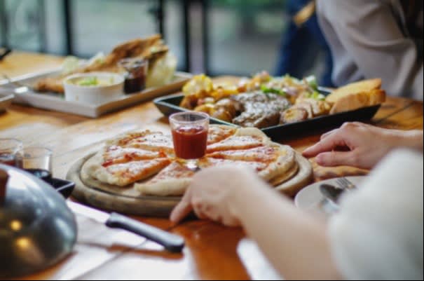 A pizza on a wooden tray with a cup of sauce in the center, surrounded by other dishes like grilled meat and bread at a group dining setting.