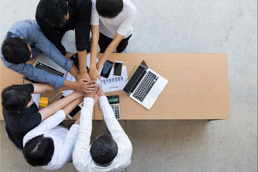 Overhead view of business team joining hands together over a desk with laptops and documents