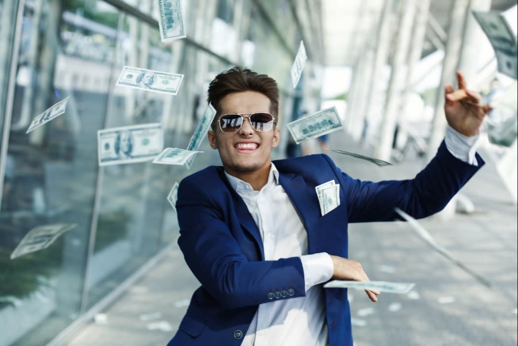 Man in a blue suit and sunglasses tossing dollar bills in the air near a modern building, symbolizing wealth, success, and financial freedom.