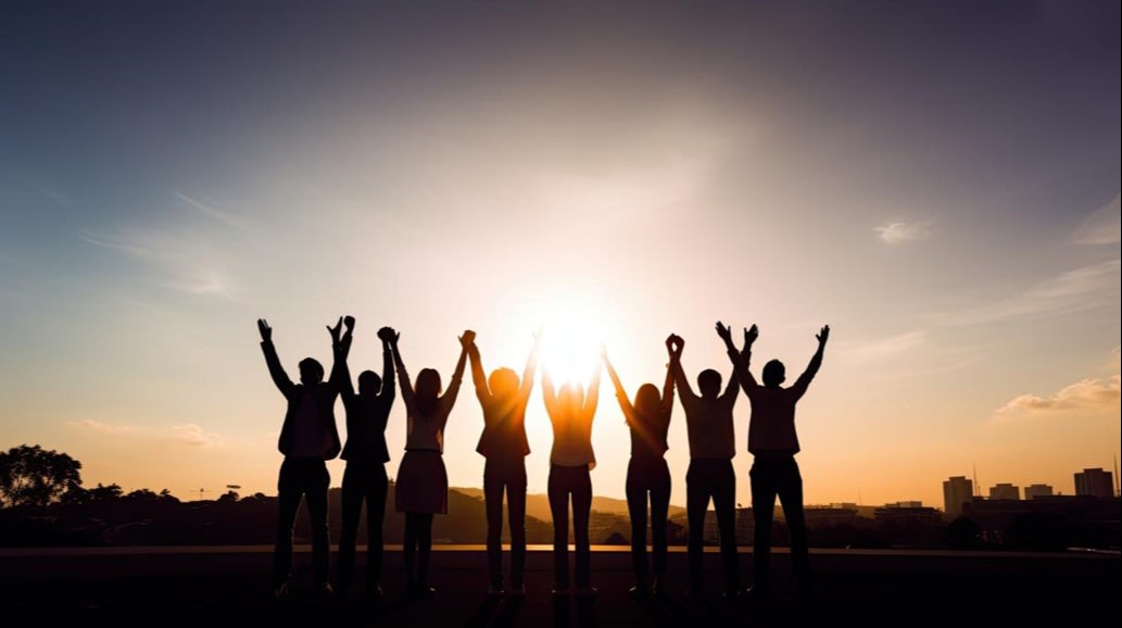 Group of people silhouetted against a sunset with arms raised, symbolizing teamwork, celebration, and unity.