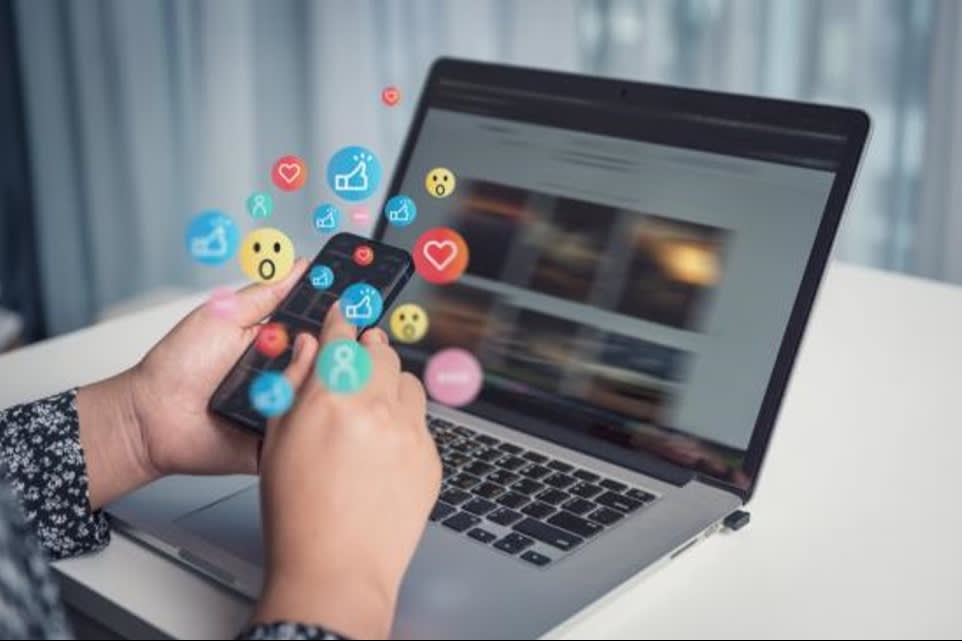 A person using a smartphone with social media reaction icons and emojis floating between the phone and laptop screen
