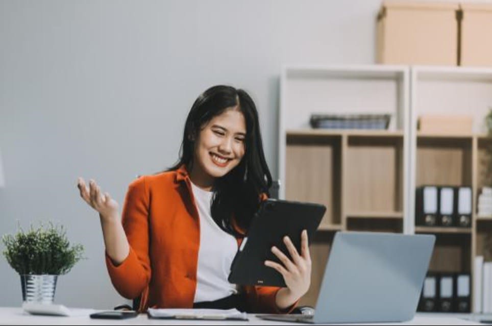 Smiling woman in a red blazer holding a tablet, working on her laptop at a modern office desk with shelves and office supplies in the background.