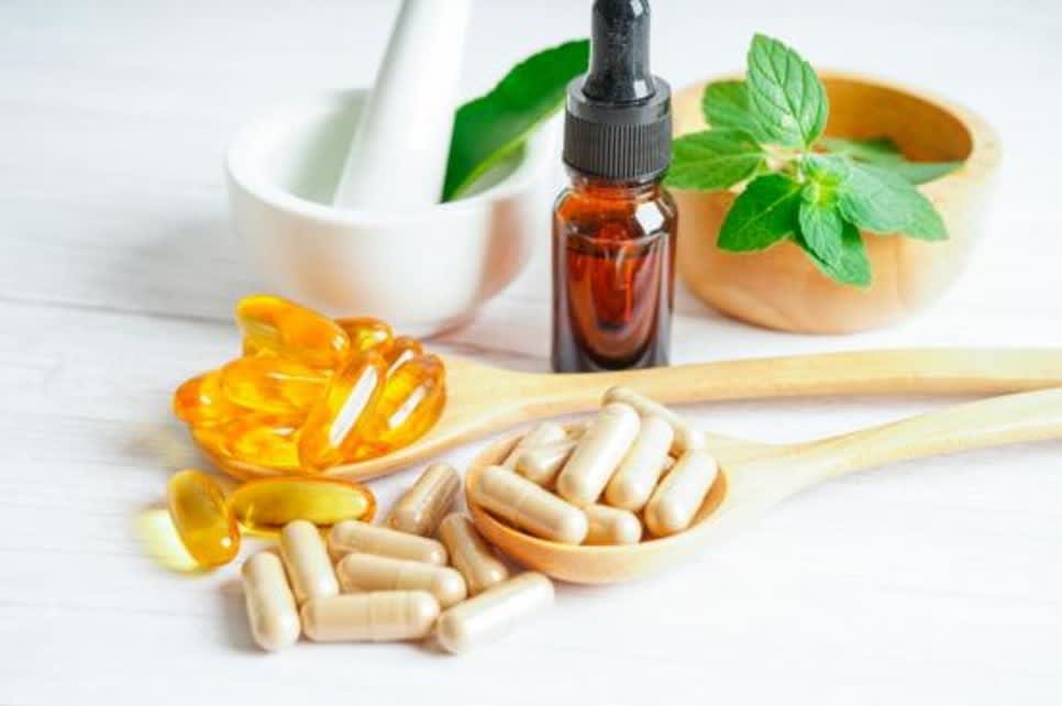 Capsules, softgels, a dropper bottle, and fresh green herbs displayed on a wooden spoon with a mortar and pestle in the background, representing natural supplements.