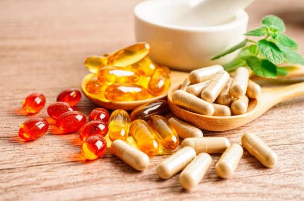 A selection of nutritional supplements, including capsules and soft gels, on a wooden surface, with green herbs and a mortar in the background.