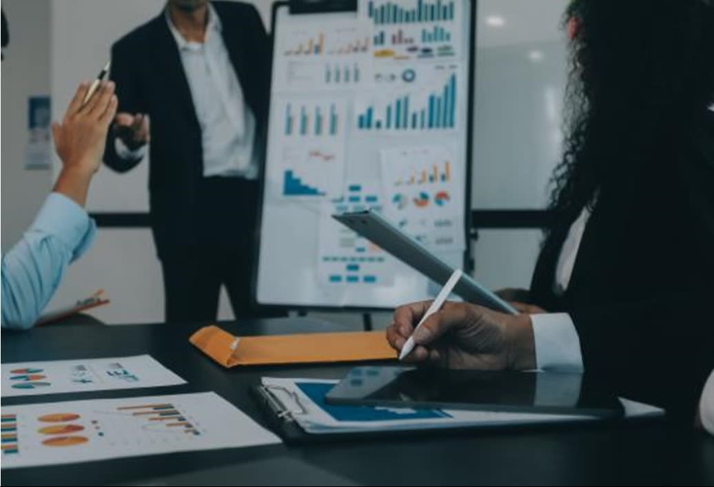 Business professionals in a meeting analyzing charts and graphs during a presentation, with one person taking notes on a tablet.