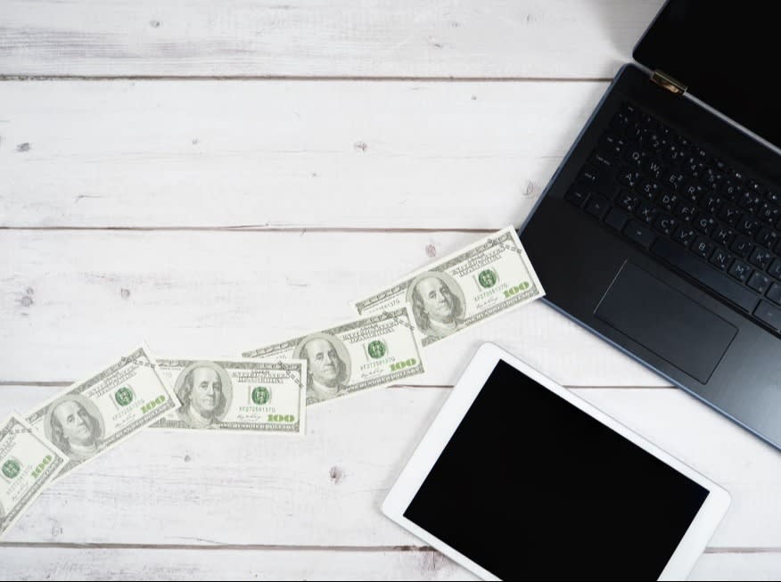 Workspace with a laptop, tablet, and cash laid out on a white wooden background.