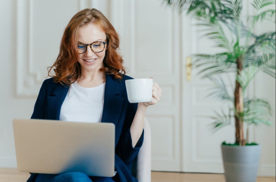 A woman with red hair and glasses smiling while working on a laptop and holding a coffee mug in a bright room with a potted plant.