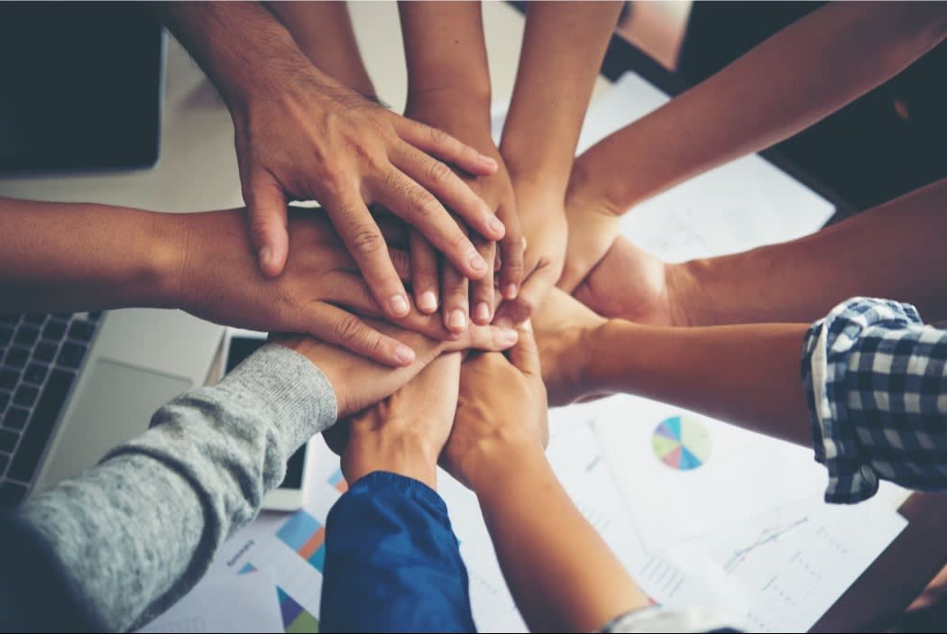 Hands stacked together in teamwork over a workspace with charts, graphs, and a laptop, symbolizing unity and collaboration.